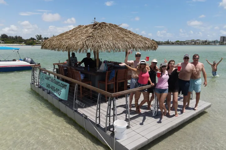 Group of people on a floating tiki bar in shallow water, holding drinks and smiling.