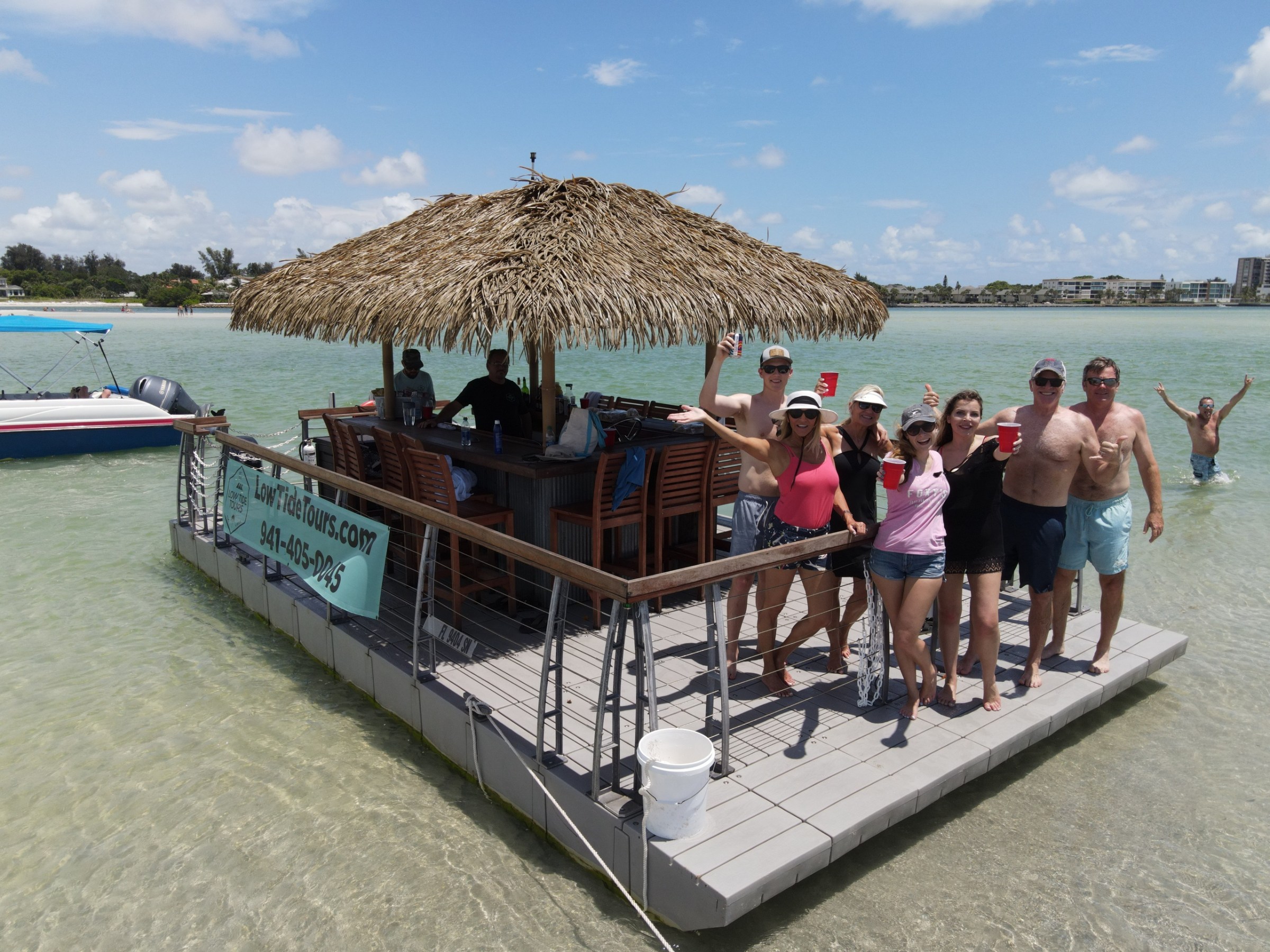 Group of people on a floating tiki bar in shallow water, holding drinks and smiling.