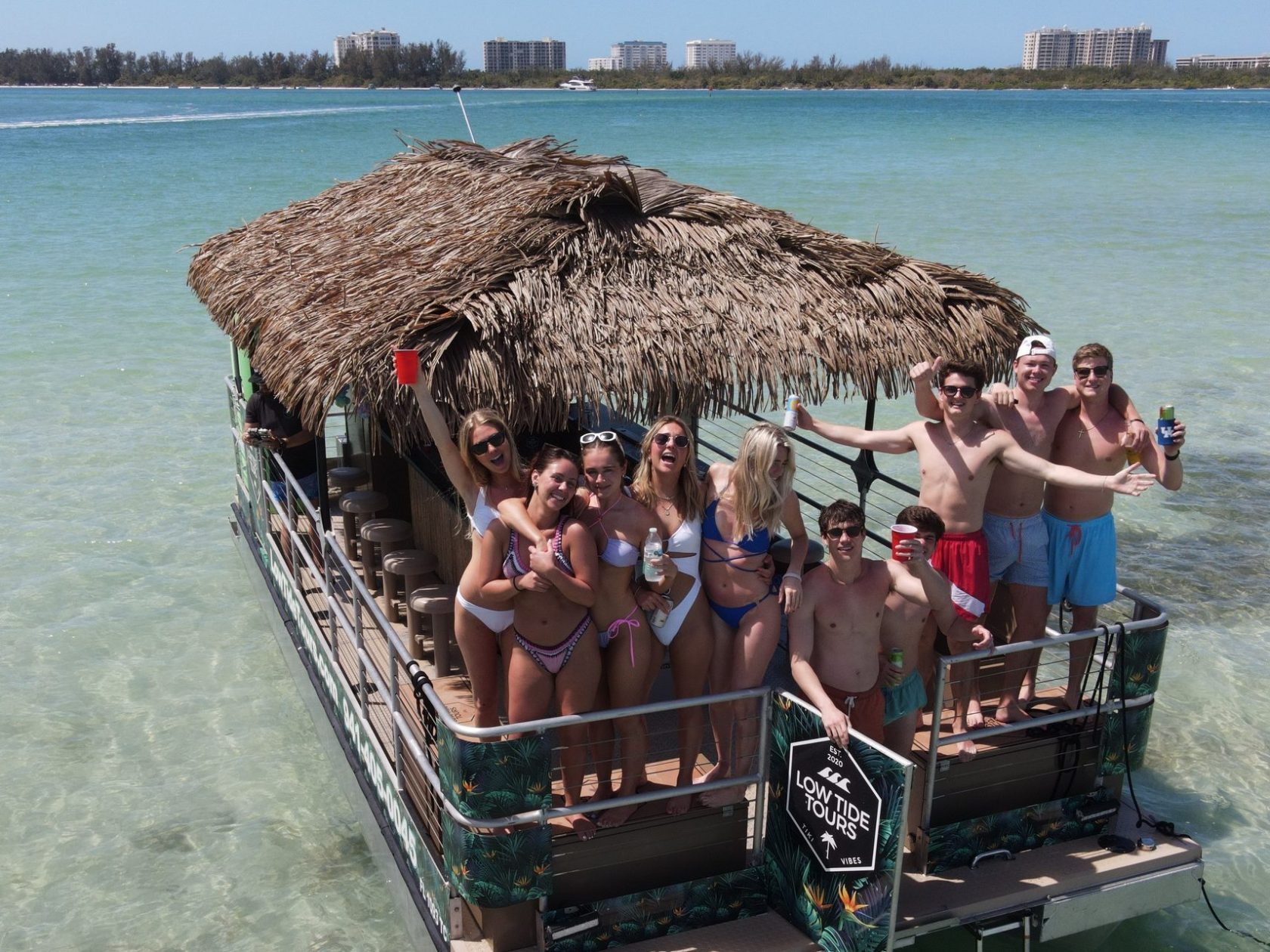 Group on a tiki boat in shallow sea, holding drinks and smiling under a clear sky.