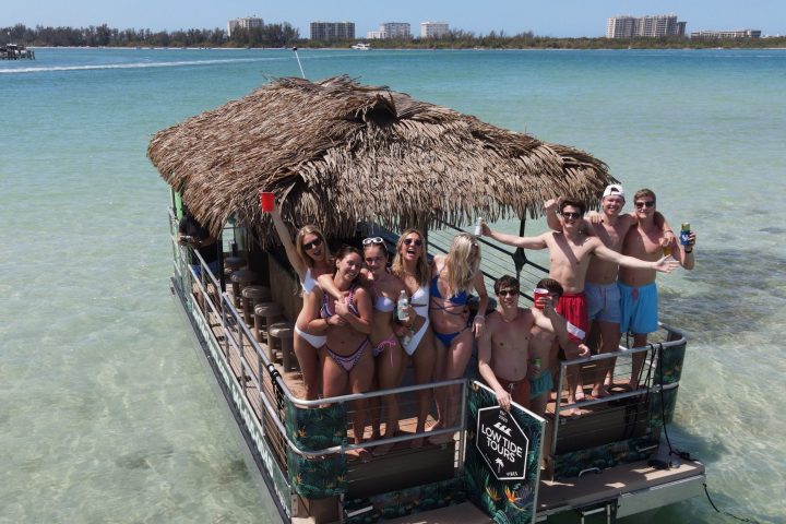 Group on a tiki boat in shallow sea, holding drinks and smiling under a clear sky.