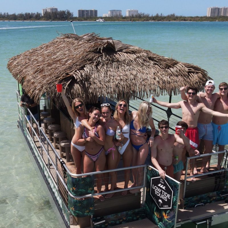 Group on a tiki boat in shallow sea, holding drinks and smiling under a clear sky.